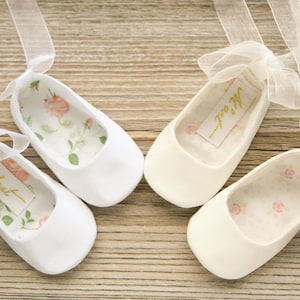 May include: Two pairs of white satin baby shoes with delicate floral lining and sheer white ribbon ties. The shoes are labeled "She's out" and "He's out".