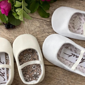 May include: Two pairs of white satin baby shoes with a floral pattern lining. One pair has the word "Matt" written on the lining.