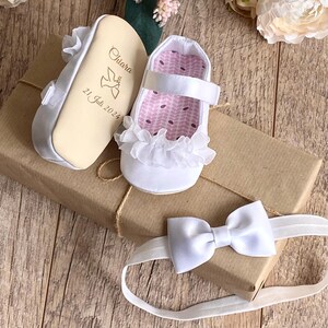 K&ouml;nnte beinhalten: Wei&szlig;e Satin-Babyschuhe mit einem wei&szlig;en R&uuml;schenbesatz aus Spitze und einer personalisierten Holzsohle. Die Sohle ist mit dem Namen Chiara und dem Datum 21. Juli 2024 graviert. Ein wei&szlig;es Satin-Haarband mit Schleife ist ebenfalls enthalten.