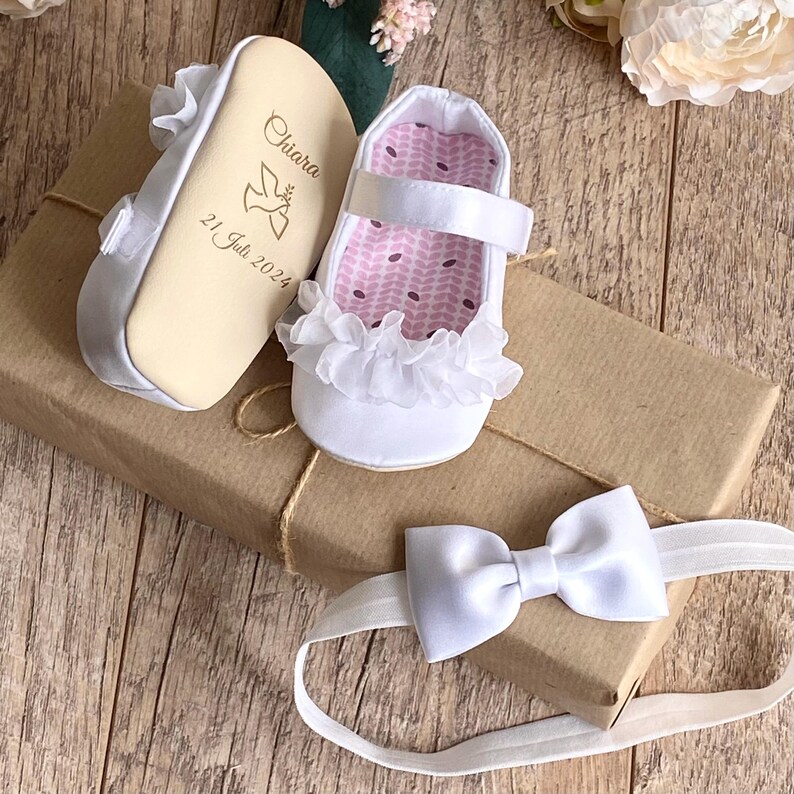 K&ouml;nnte beinhalten: Wei&szlig;e Satin-Babyschuhe mit einem wei&szlig;en R&uuml;schenbesatz aus Spitze und einer personalisierten Holzsohle. Die Sohle ist mit dem Namen Chiara und dem Datum 21. Juli 2024 graviert. Ein wei&szlig;es Satin-Haarband mit Schleife ist ebenfalls enthalten.