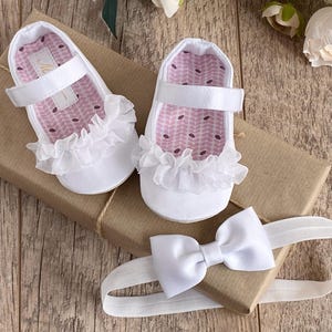 K&ouml;nnte beinhalten: Wei&szlig;e Babyschuhe mit R&uuml;schenbesatz und einem wei&szlig;en Satin-Haarband mit Schleife. Die Schuhe haben ein rosa-wei&szlig;es gemustertes Futter.