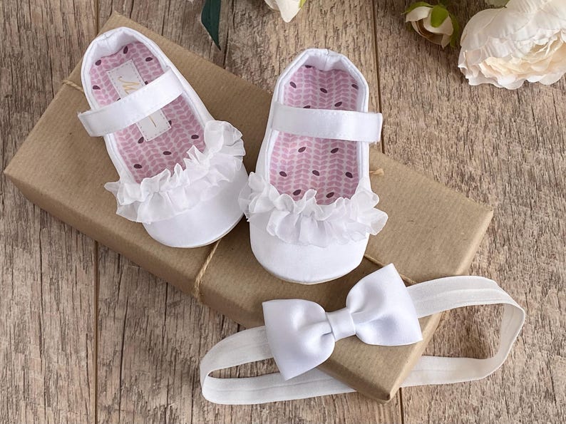 K&ouml;nnte beinhalten: Wei&szlig;e Babyschuhe mit R&uuml;schenbesatz und einem wei&szlig;en Satin-Haarband mit Schleife. Die Schuhe haben ein rosa-wei&szlig;es gemustertes Futter.