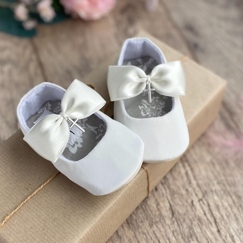 May include: White satin baby shoes with a satin bow and a silver cross charm. The shoes are on a brown gift box.