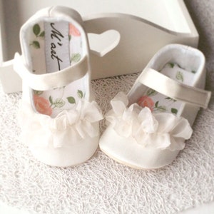 K&ouml;nnte beinhalten: Ein Paar wei&szlig;e Satin-Babyschuhe mit R&uuml;schenbesatz und einem floralen Futter. Die Schuhe haben einen Riemen &uuml;ber dem Oberteil und sagen "Mi'aet" auf der Innenseite.