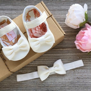 May include: A set of white baby shoes with a bow on the front, a white headband with a bow, and two pink and white peonies.