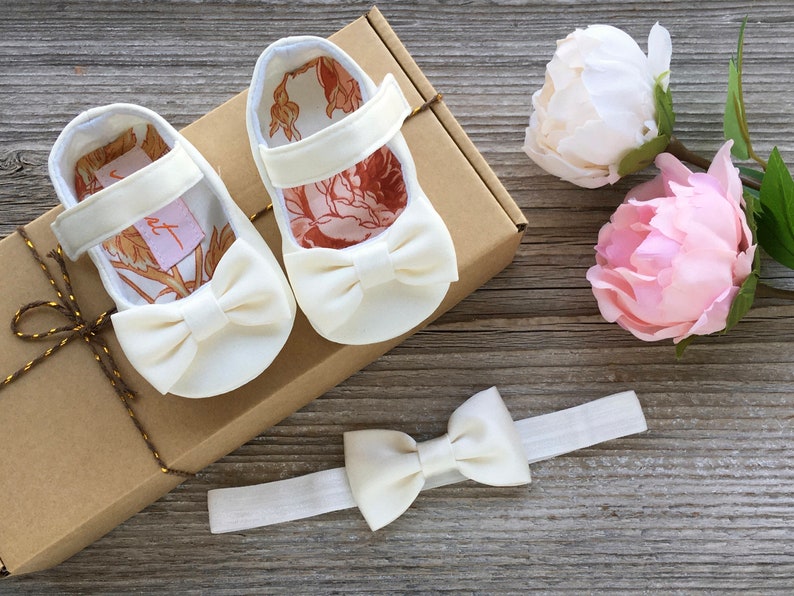 May include: A set of white baby shoes with a bow on the front, a white headband with a bow, and two pink and white peonies.