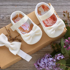 May include: A set of white baby shoes with a bow and a matching white headband with a bow. The shoes and headband are in a brown box with a gold ribbon.
