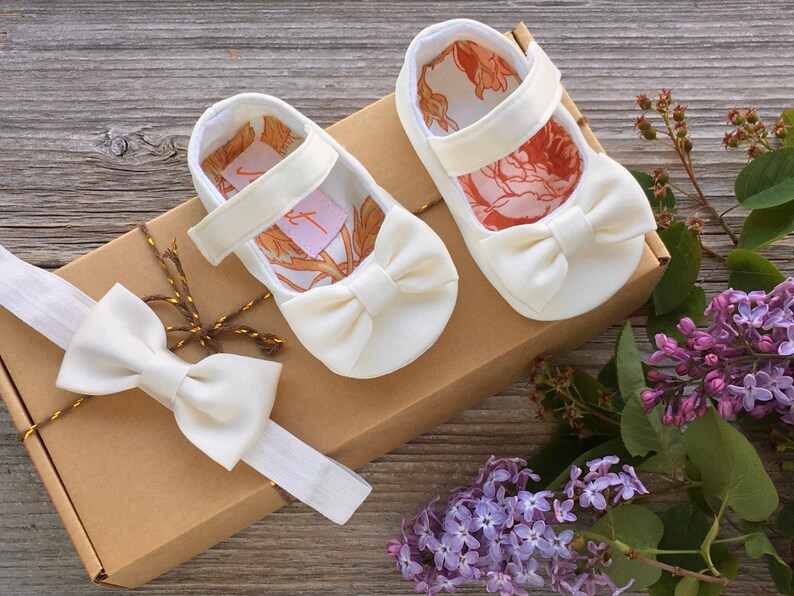 May include: A set of white baby shoes with a bow and a matching white headband with a bow. The shoes and headband are in a brown box with a gold ribbon.