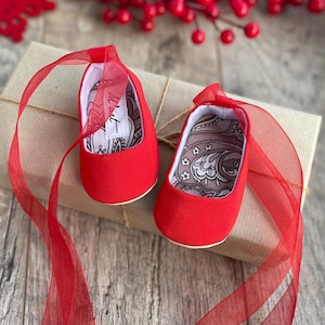 Könnte beinhalten: Ein Paar rote Babyschuhe mit Paisley-Musterfutter und roten Satinbändern. Die Schuhe befinden sich auf einem braunen Geschenkpaket.