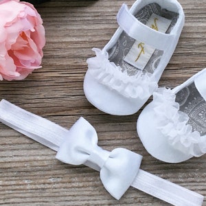 K&ouml;nnte beinhalten: Wei&szlig;e Babyschuhe mit einem wei&szlig;en Schleifen-Haarband. Die Schuhe haben einen wei&szlig;en Spitzenbesatz und ein graues Paisley-Muster innen. Das Haarband hat eine wei&szlig;e Schleife aus Satin.