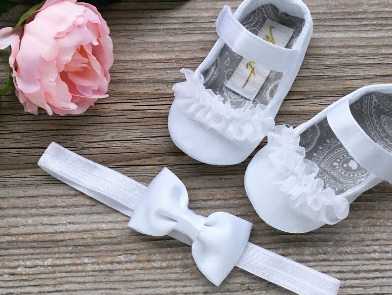 K&ouml;nnte beinhalten: Wei&szlig;e Babyschuhe mit einem wei&szlig;en Schleifen-Haarband. Die Schuhe haben einen wei&szlig;en Spitzenbesatz und ein graues Paisley-Muster innen. Das Haarband hat eine wei&szlig;e Schleife aus Satin.