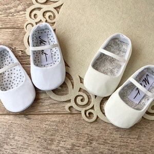 May include: Three pairs of white baby shoes with a satin finish. The shoes have a simple design with a single strap across the top. The lining of the shoes is white with a pattern of small grey dots or a floral design.
