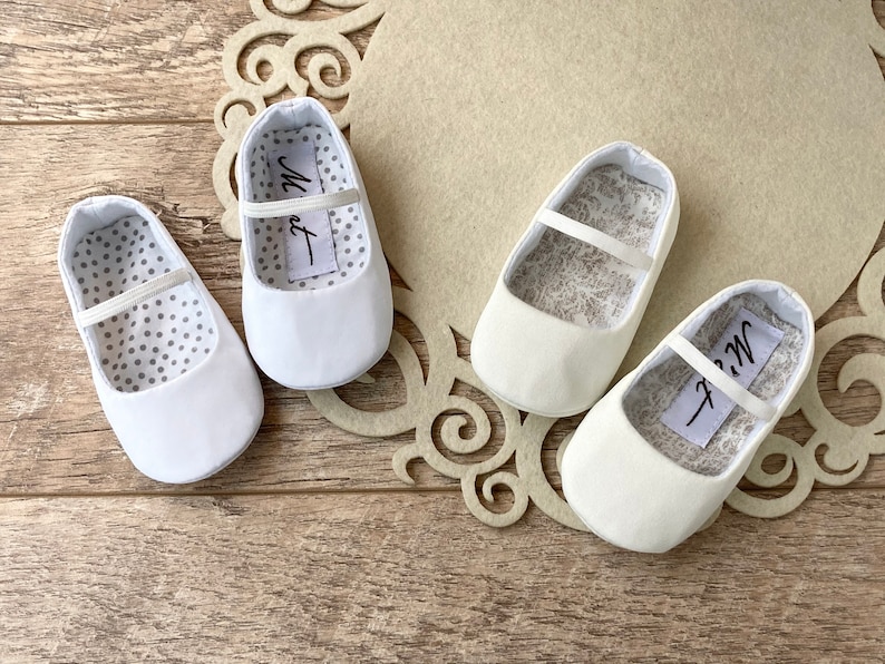 May include: Three pairs of white baby shoes with a satin finish. The shoes have a simple design with a single strap across the top. The lining of the shoes is white with a pattern of small grey dots or a floral design.
