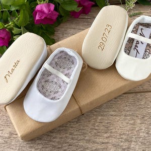 May include: Three pairs of white baby shoes with different inscriptions on the soles. The shoes are on a brown box with a white ribbon. The inscriptions on the soles are "Marta", "29.07.23", and "Nora".