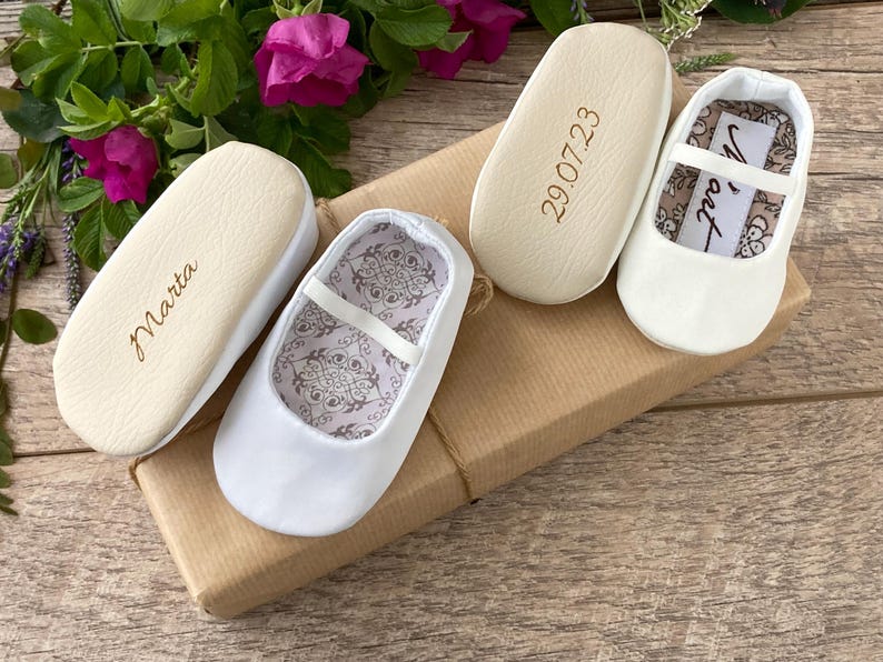 May include: Three pairs of white baby shoes with different inscriptions on the soles. The shoes are on a brown box with a white ribbon. The inscriptions on the soles are "Marta", "29.07.23", and "Nora".