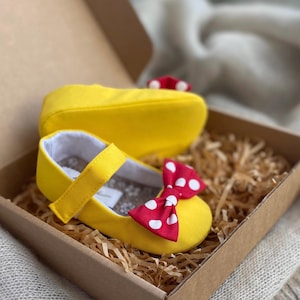 K&ouml;nnte beinhalten: Ein Paar gelbe Babyschuhe mit roten Tupfen-Schleifen. Die Schuhe befinden sich in einem braunen Karton mit zerkn&uuml;lltem Papier.