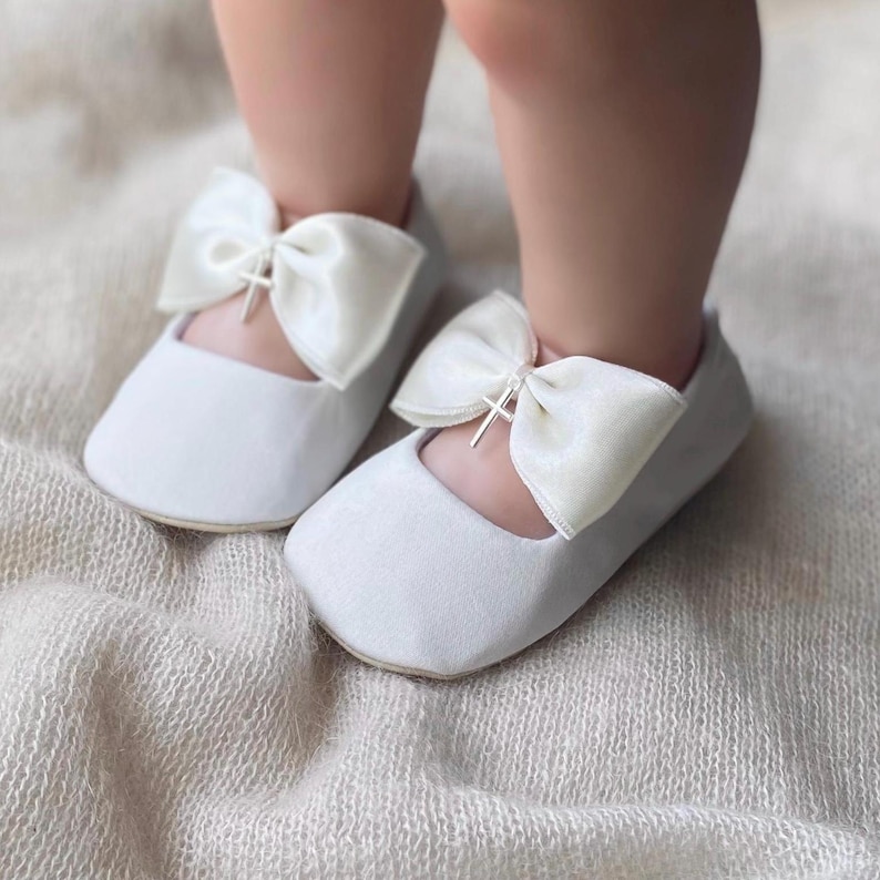 May include: White satin baby shoes with a satin bow and a silver cross accent. The shoes are on a light brown fuzzy surface.