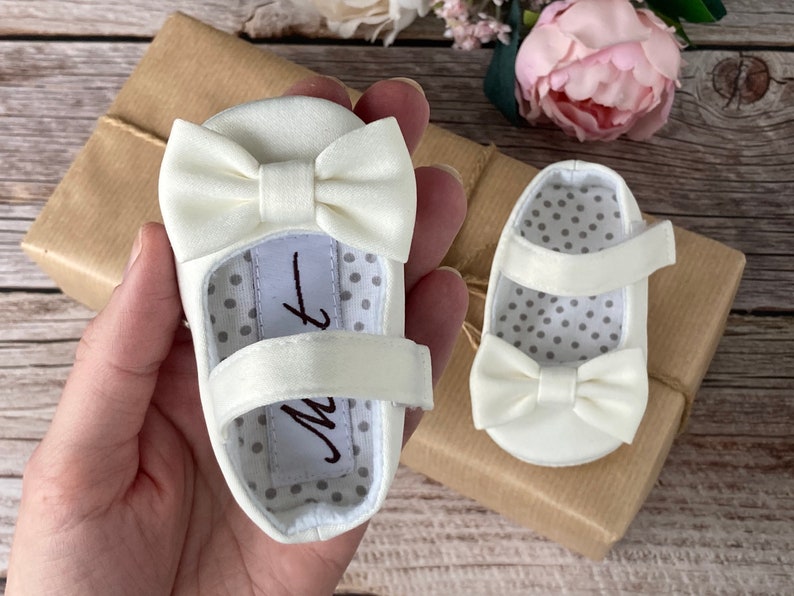 May include: A pair of white satin baby shoes with a bow on the toe and a strap across the foot. The shoes have a white polka dot lining.