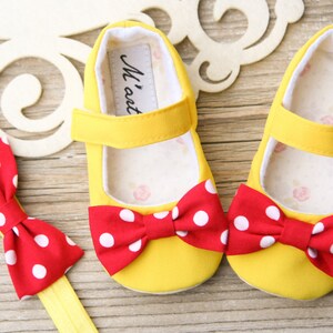 K&ouml;nnte beinhalten: Ein Paar gelbe Babyschuhe mit roten und wei&szlig;en Tupfen-Schleifen. Die Schuhe haben einen Klettverschluss. Zu den Schuhen geh&ouml;rt ein passendes Stirnband mit einer roten und wei&szlig;en Tupfen-Schleife.