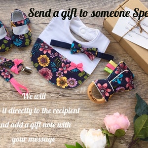 K&ouml;nnte beinhalten: Ein Baby-Geschenkset mit einer floralen L&auml;tzchen, einer Fliege, Schuhen und einem Holz-Bei&szlig;ring mit dem eingravierten Namen "Emma". Das Set ist in einer braunen Box mit einem Geschenkanh&auml;nger verpackt.