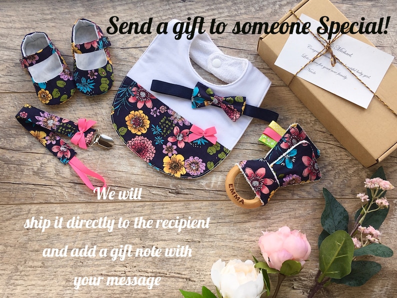 K&ouml;nnte beinhalten: Ein Baby-Geschenkset mit einer floralen L&auml;tzchen, einer Fliege, Schuhen und einem Holz-Bei&szlig;ring mit dem eingravierten Namen "Emma". Das Set ist in einer braunen Box mit einem Geschenkanh&auml;nger verpackt.