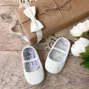 May include: A pair of white satin baby shoes with a small white bow on each shoe. The shoes are on a wooden surface with a brown gift box and white flowers.