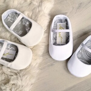 May include: A pair of white baby shoes with a gray and white patterned lining. The shoes have a simple design with a single strap across the top. The lining of the shoes has the word "Meet" written on it.