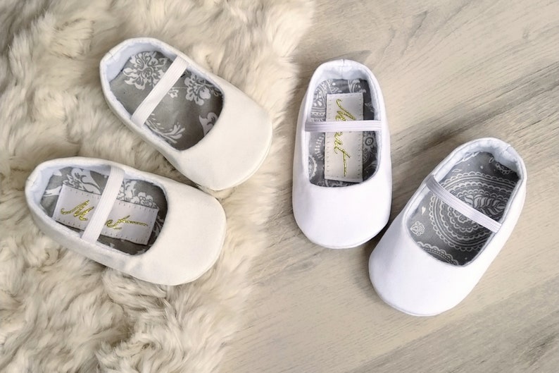 May include: A pair of white baby shoes with a gray and white patterned lining. The shoes have a simple design with a single strap across the top. The lining of the shoes has the word "Meet" written on it.
