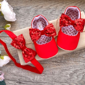 Peut inclure: Chaussures de bébé rouges avec un nœud en paillettes et un bandeau rouge assorti avec un nœud en paillettes. Les chaussures et le bandeau sont sur une boîte cadeau marron.