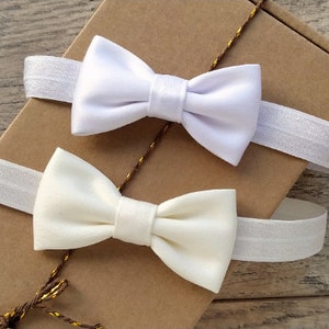 May include: Two white satin bow ties with elastic bands. The bow ties are on a brown paper background.