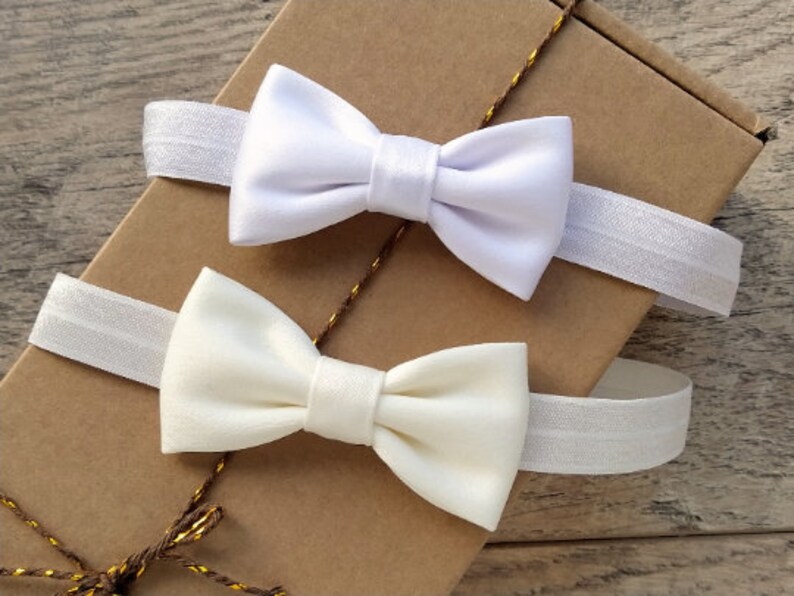 May include: Two white satin bow ties with elastic bands. The bow ties are on a brown paper background.