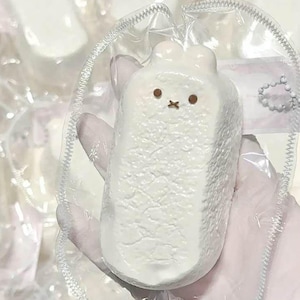 May include: A white, rectangular, textured figurine shaped like a rabbit with two small ears at the top. The face features two brown circular eyes and a brown cross-shaped mouth. The item is encased in a clear, crinkled plastic packaging.