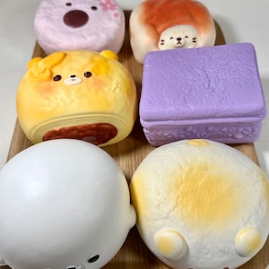 May include: A collection of six soft, squishy foam toys shaped like various foods and animals. The set includes a pink donut, a bear-shaped bread roll, a yellow bear bun, a purple rectangular block, a white seal, and a round white bun with ears.