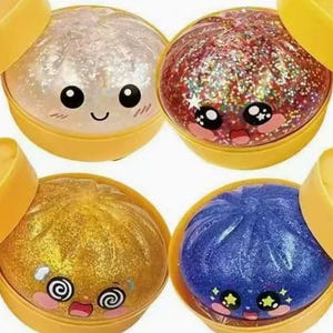 May include: Four colorful, glitter-filled toy dumplings in yellow containers. The dumplings are white, red, gold, and blue, each with a different kawaii face. The containers have a lid that is partially open.