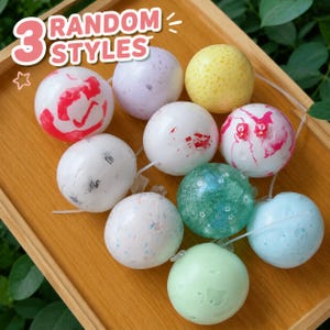 May include: A collection of bath bombs in various colors and patterns, including red, white, yellow, green, and blue. The image includes the text "3 RANDOM STYLES" in a playful font, suggesting a variety of options.