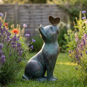 May include: A bronze-colored cat statue with a butterfly perched on its head, set in a garden. The cat is in a seated position, gazing upwards. Lavender and other flowers surround the statue, creating a serene outdoor scene.