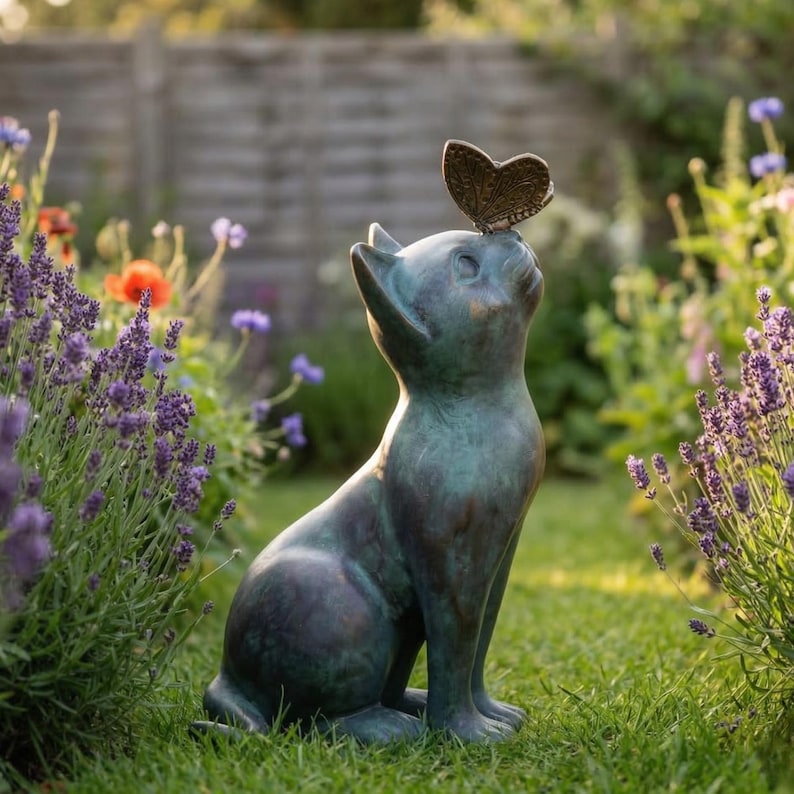 May include: A bronze-colored cat statue with a butterfly perched on its head, set in a garden. The cat is in a seated position, gazing upwards. Lavender and other flowers surround the statue, creating a serene outdoor scene.