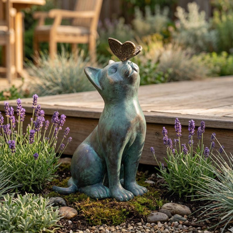 May include: A bronze-colored cat statue with a butterfly perched on its nose. The cat is sitting upright, gazing upwards. The statue is placed outdoors, surrounded by lavender plants and a wooden deck.