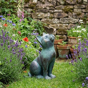 May include: A verdigris-colored cat statue with a butterfly perched on its nose, set in a lush garden. The cat is gazing upwards, with a detailed butterfly. The garden features colorful flowers and a stone wall backdrop.