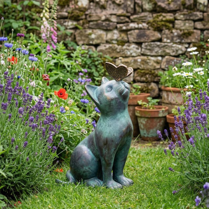 May include: A verdigris-colored cat statue with a butterfly perched on its nose, set in a lush garden. The cat is gazing upwards, with a detailed butterfly. The garden features colorful flowers and a stone wall backdrop.