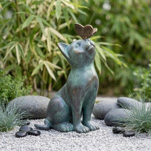 May include: A bronze-colored cat statue with a butterfly perched on its head. The cat is sitting upright, looking up with an expression of curiosity. The statue has a verdigris finish, suggesting it is made of aged metal. The statue is placed on a bed of small gray stones.