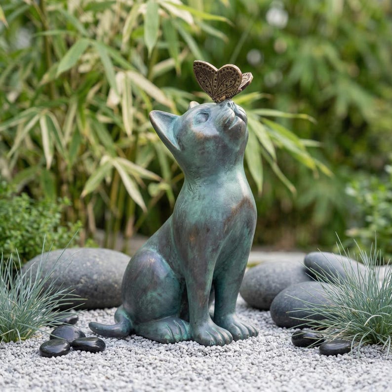 May include: A bronze-colored cat statue with a butterfly perched on its head. The cat is sitting upright, looking up with an expression of curiosity. The statue has a verdigris finish, suggesting it is made of aged metal. The statue is placed on a bed of small gray stones.