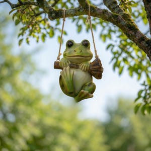 May include: A whimsical green frog figurine on a swing made of rope and a wooden perch. The frog has large, expressive eyes and is suspended from a tree branch. The background is blurred with green foliage and a hint of blue sky.