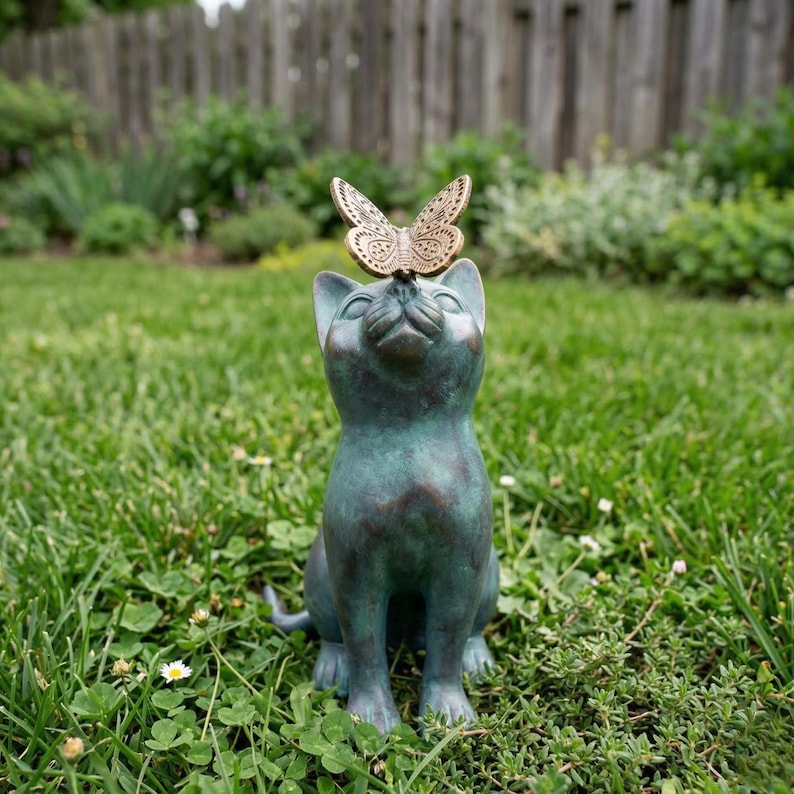 May include: A bronze-colored cat statue with a butterfly perched on its head, set in a grassy outdoor scene. The cat is looking upwards with a serene expression. The butterfly has intricate wing details.