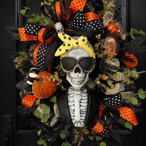 Può includere: Una ghirlanda di Halloween con un teschio che indossa occhiali da sole e un foulard giallo a pois. La ghirlanda include nastri a pois arancioni e neri, piccole zucche e fiori neri. La ghirlanda è esposta su una porta nera.