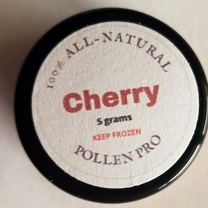 May include: A round, black container with a white label. The label features the word "cherry" in red, along with "5 grams" and "100% ALL-NATURAL." The text "KEEP FROZEN" and "POLLEN PRO" are also visible.