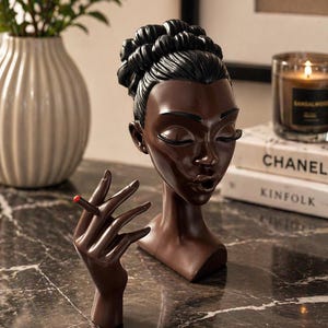 May include: A brown resin sculpture of a woman's head and hand holding a cigarette with a red tip. The woman's hair is styled in a bun. The sculpture is on a marble surface.