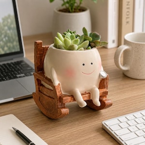 Op de afbeelding: Een grillige, crèmekleurige keramische plantenbak in de vorm van een lachend karakter dat in een houten schommelstoel zit. De plantenbak is gevuld met groene vetplanten. Een decoratief item voor thuis of op kantoor.