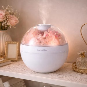 May include: A white, spherical humidifier with a clear dome filled with pink flowers. The word "humidifier" is printed on the base. A stream of vapour rises from the top. The humidifier sits on a white shelf with other decorative items.