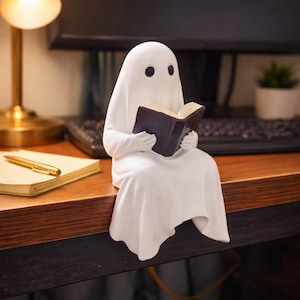 Op de afbeelding: Een wit spookbeeldje dat een boek met een donkerblauwe kaft leest. Het spook heeft twee zwarte oog gaten en zit. Het beeldje staat op een houten bureau, met een lamp en een notitieboek op de achtergrond.